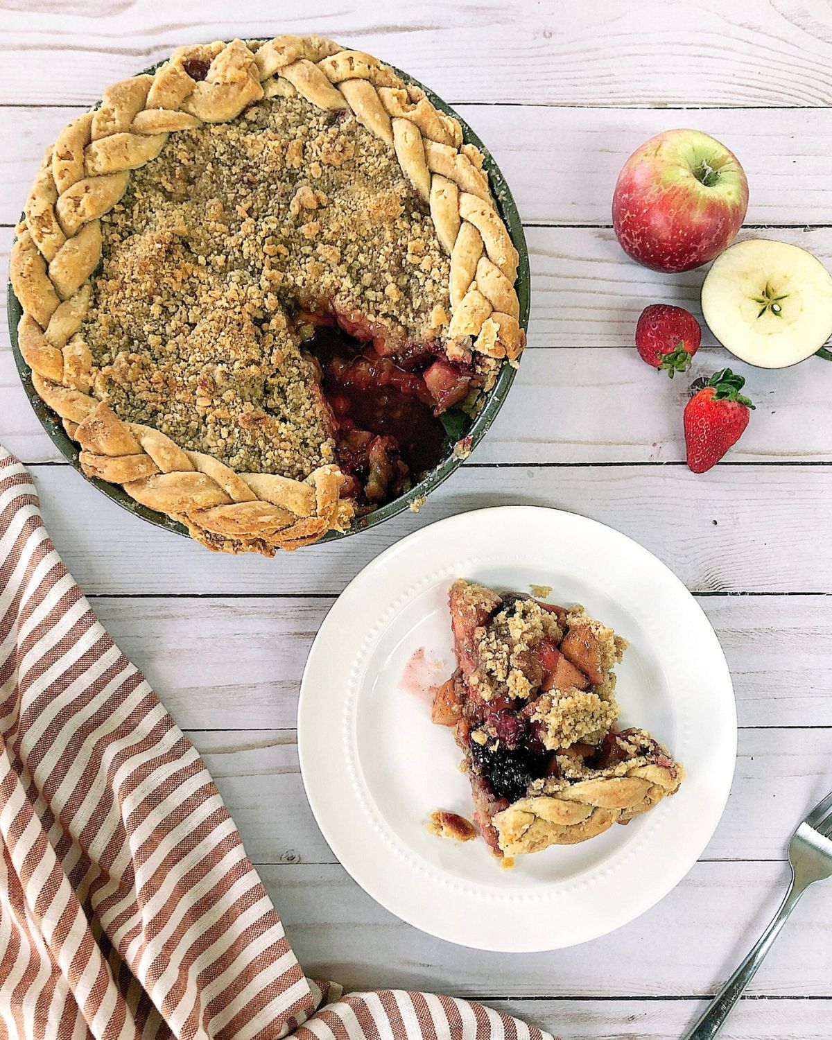 Apple mountain berry crumb pie with a braided crust edge and golden crumb topping, sliced to show berry and apple filling, with a slice plated beside fresh apples and strawberries