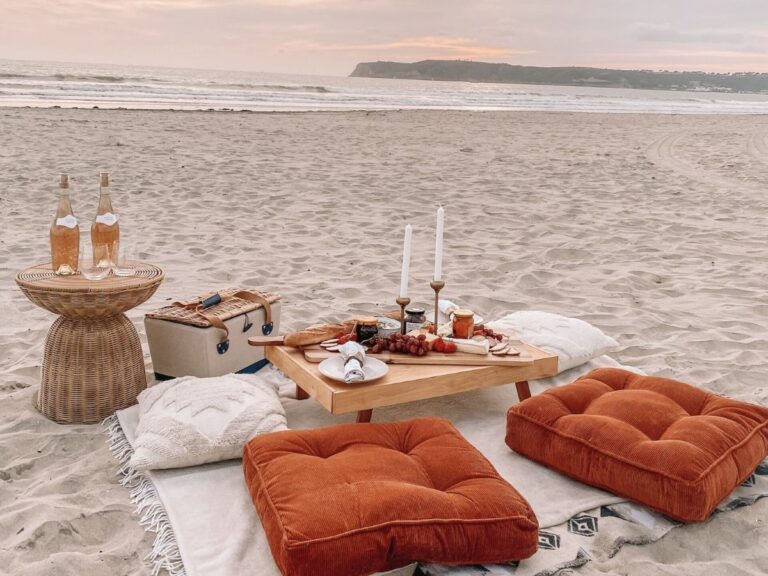 Wide-angle view of a complete beach picnic setup at sunset on Coronado — wicker side table, low wooden table with charcuterie board and candles, burnt orange floor pillows, and the Pacific in the distance.