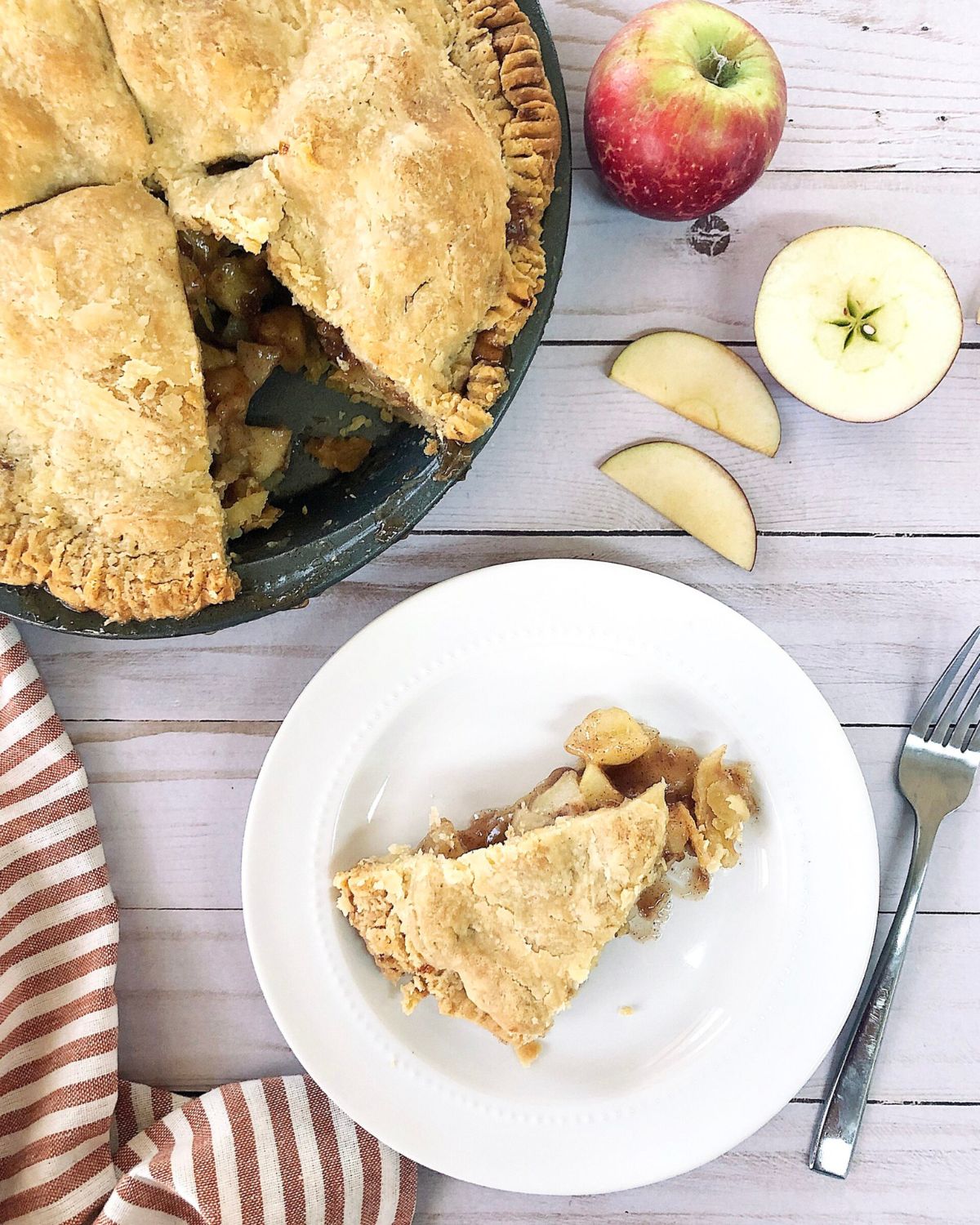 Homemade apple pie with a golden brown crimped crust and vent slits, one slice removed and plated to show the cinnamon apple filling, with fresh apples styled beside it on a white wood surface