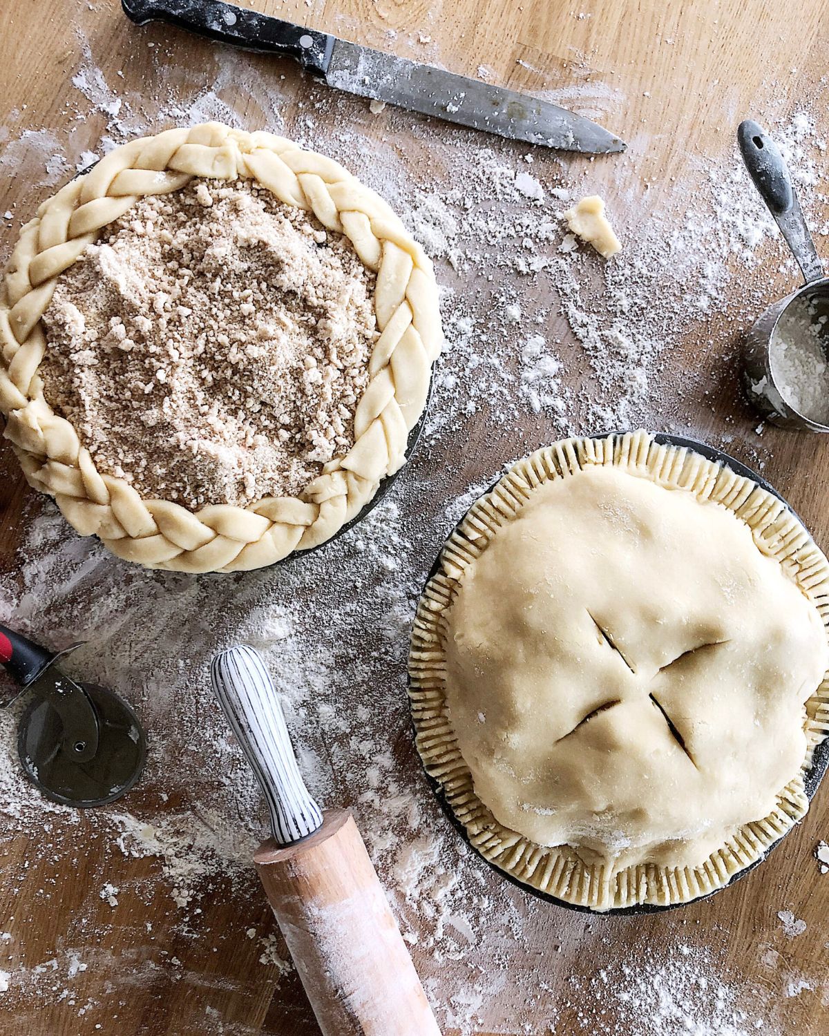 Two unbaked homemade pie crusts on a flour-dusted wooden surface — one with a braided edge topped with brown sugar crumb, one fully covered with a fork-crimped edge and vent slits