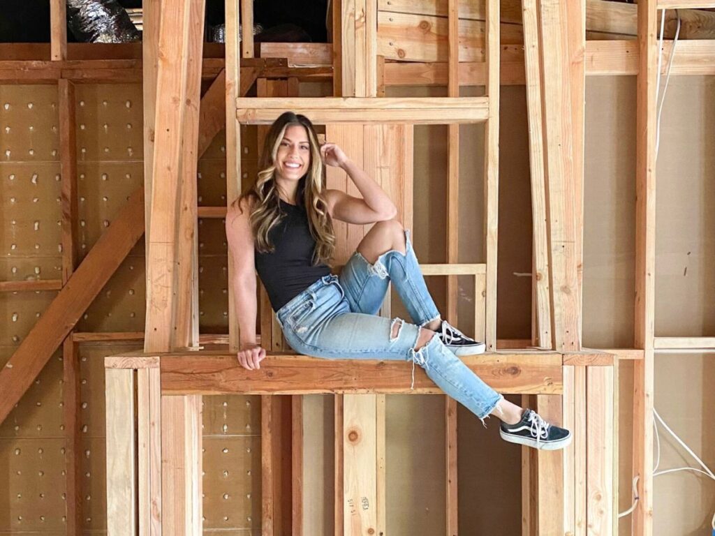 A woman sitting on exposed wood framing inside a home mid-renovation — the structural phase where renovation budgets are most often tested.