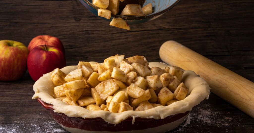 Homemade apple pie filling in an unbaked crust on a rustic wood surface with fresh apples and a wooden rolling pin