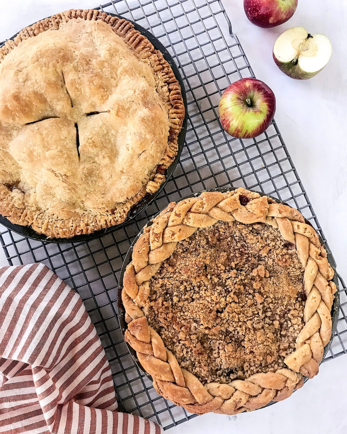 Two homemade pies cooling on a black wire rack &mdash; a classic apple pie with a vented golden crust and an apple mountain berry crumb pie with a braided crust edge