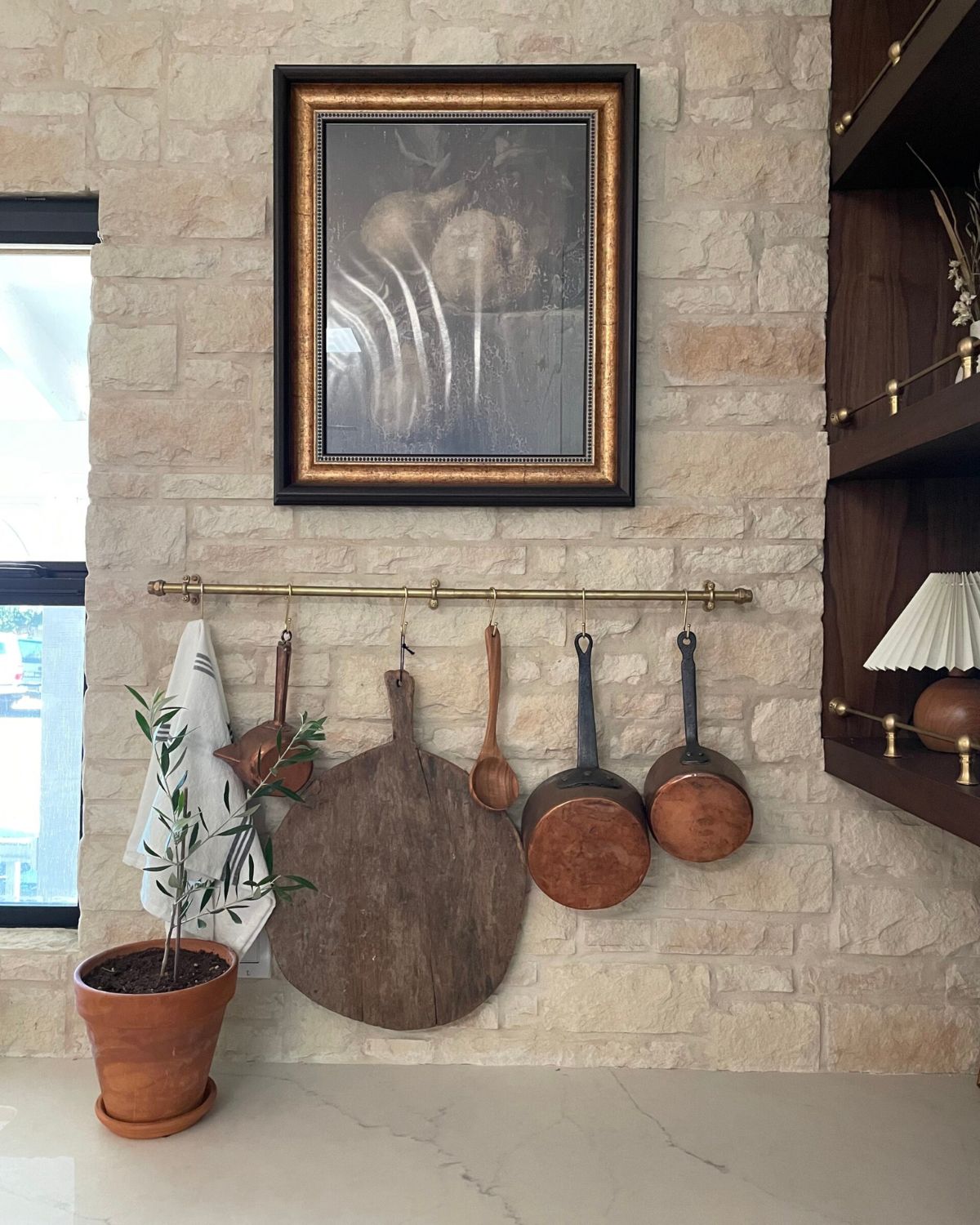 Tall gilt-framed art hung above a brass pot rail with copper pans on a stone kitchen wall — interior design scale and proportion example
