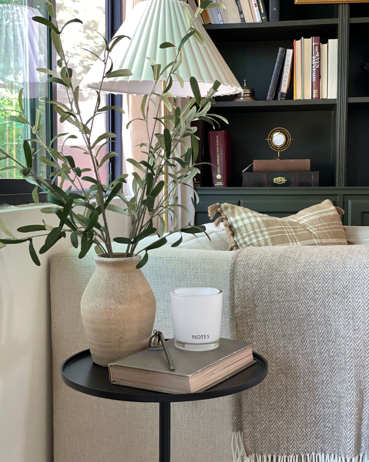 Black round side table styled with ceramic vase, olive branches, antique book, and Notes candle — rule of three interior design tip for beginners