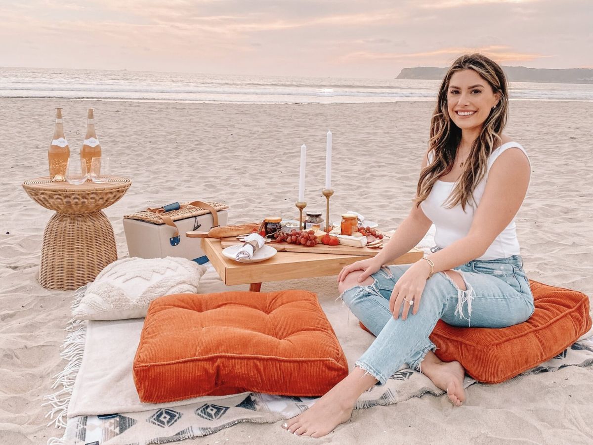 Liz Lovery styling a beach picnic on Coronado at sunset — cream blanket, low wooden table with charcuterie, wicker side table with rosé, and burnt-orange floor pillows on the sand