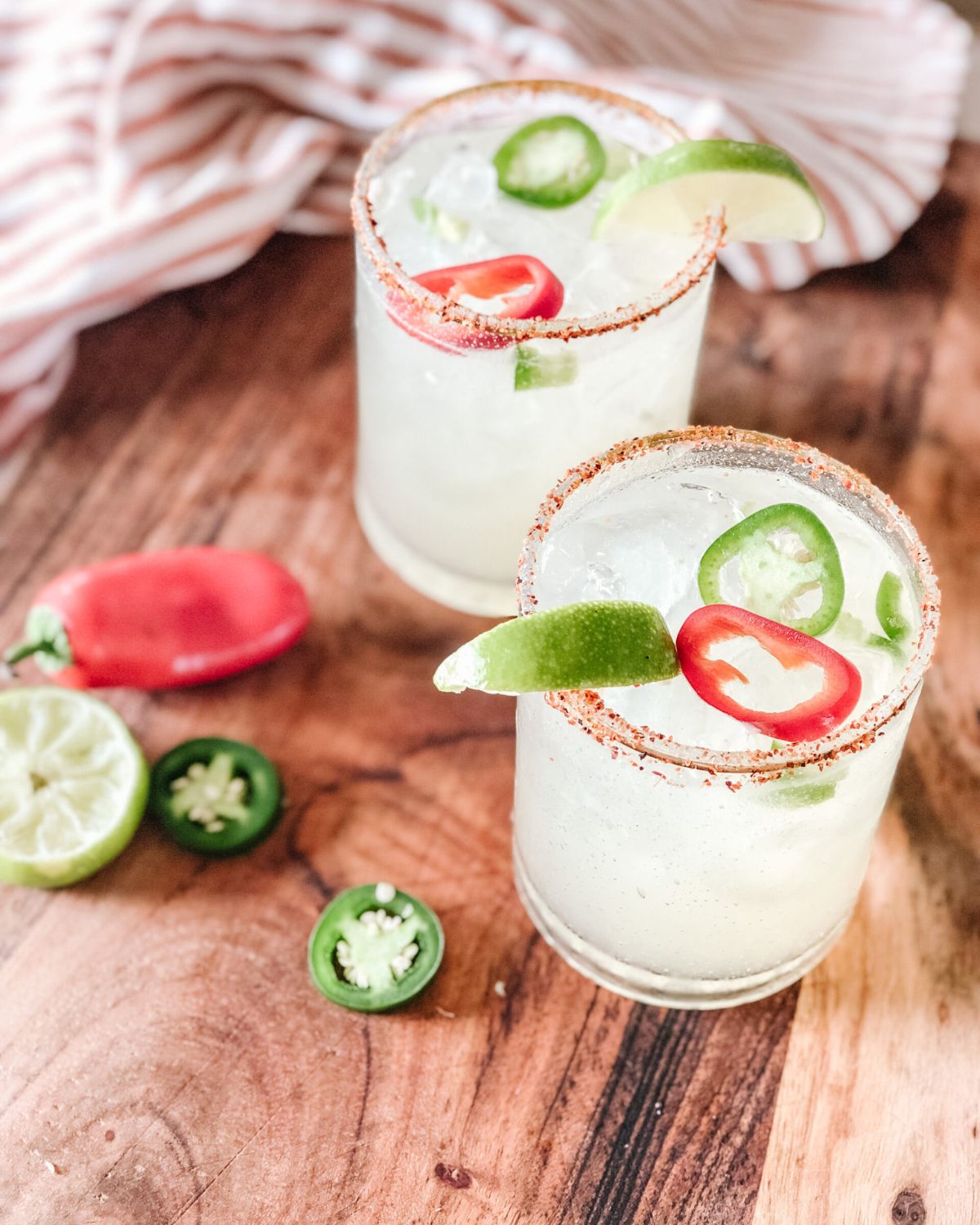 Spicy margaritas garnished with fresh jalapeño slices, lime wedges, and red chili pepper rounds on a wooden board.