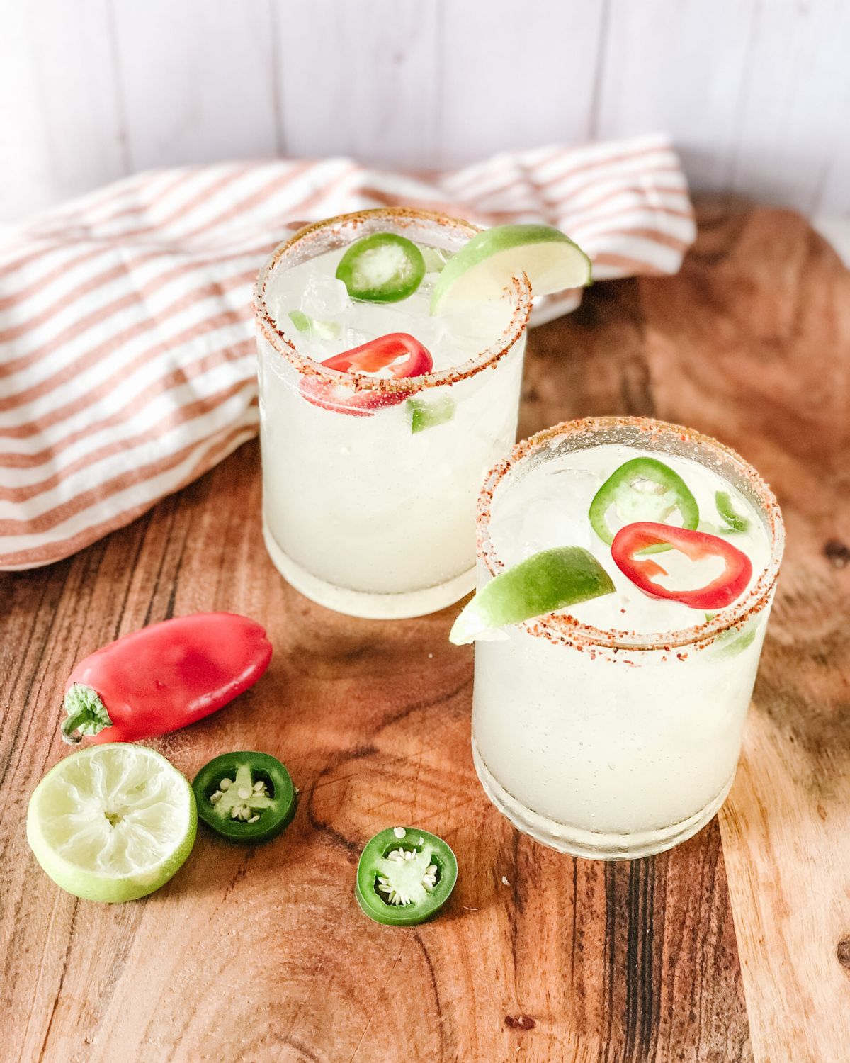 Two spicy margaritas with salt rims, jalapeño slices, lime wedges, and chili pepper garnish on a wooden board.