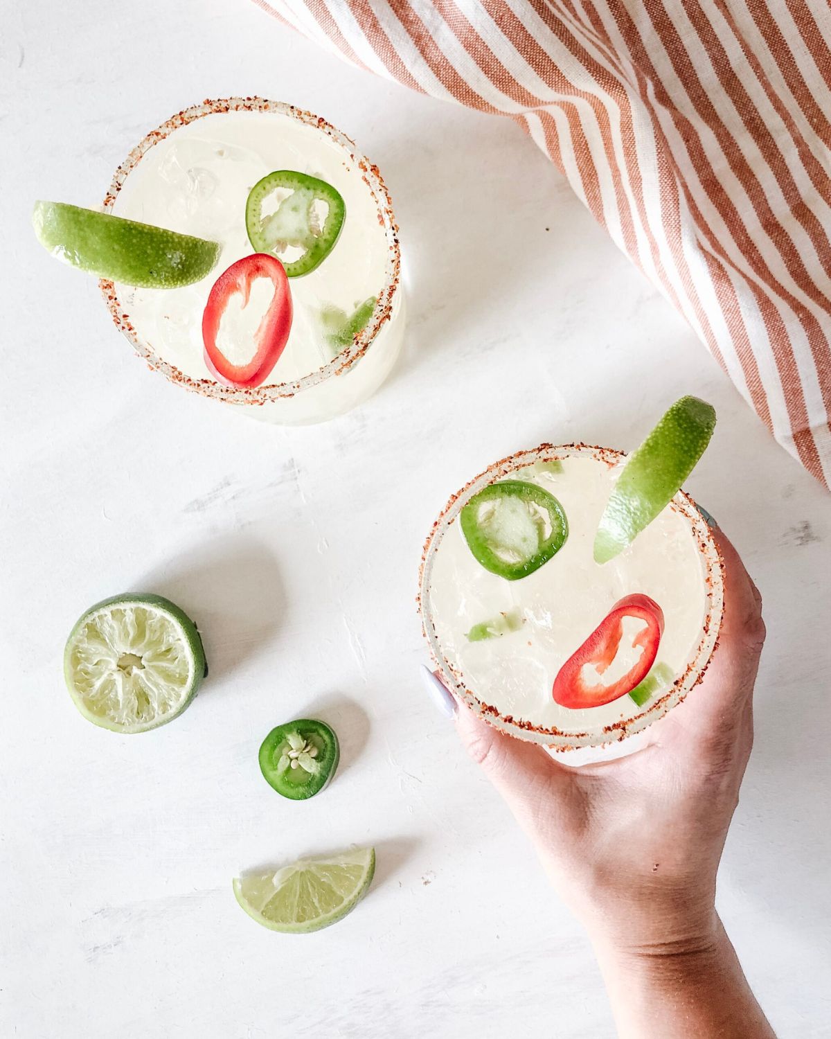 Overhead view of a hand holding a salt-rimmed spicy margarita with jalapeño and chili pepper garnish, halved lime nearby.