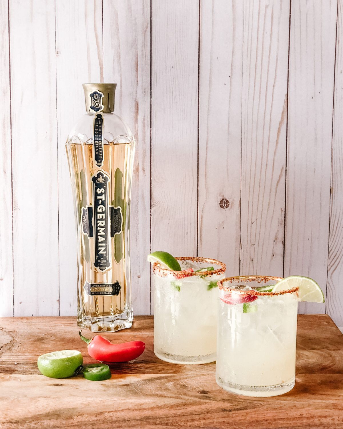 St. Germain elderflower liqueur bottle on a wooden board next to two spicy margaritas with salt rims, lime, and chili pepper garnish.