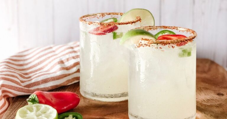 Two spicy margaritas with salt-rimmed glasses, jalapeño slices, lime wedges, and a chili pepper on a wooden board.