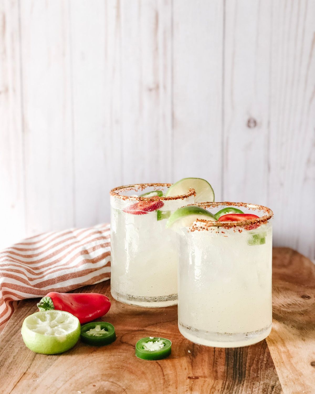 Two spicy margaritas in salt-rimmed glasses on a wooden board with a striped napkin, fresh lime, jalapeño slices, and a red chili pepper.