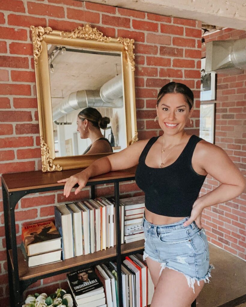 Liz Lovery posing next to her finished DIY Anthropologie mirror dupe with gold ornate frame against a brick wall