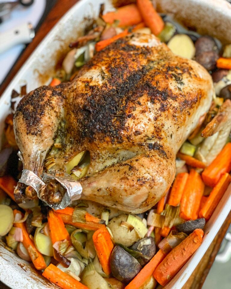 Golden roast chicken in a white pan surrounded by carrots, leeks, and potatoes