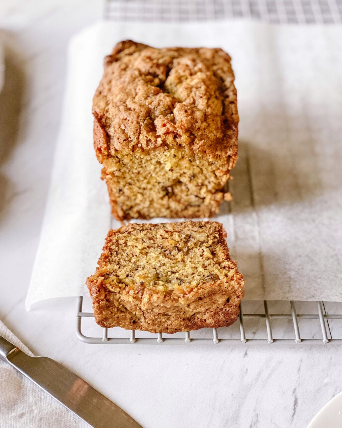 Two super moist banana bread loaves with cinnamon streusel tops cooling on a wire rack, one sliced to show the soft crumb