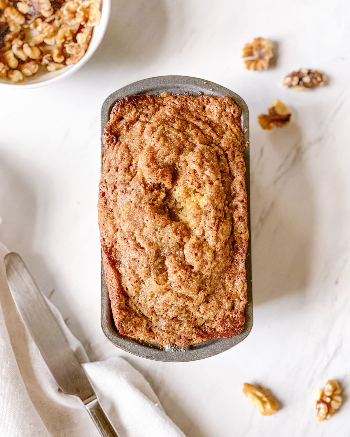 Overhead view of a super moist banana bread loaf with golden cinnamon streusel topping in a loaf pan, surrounded by walnuts