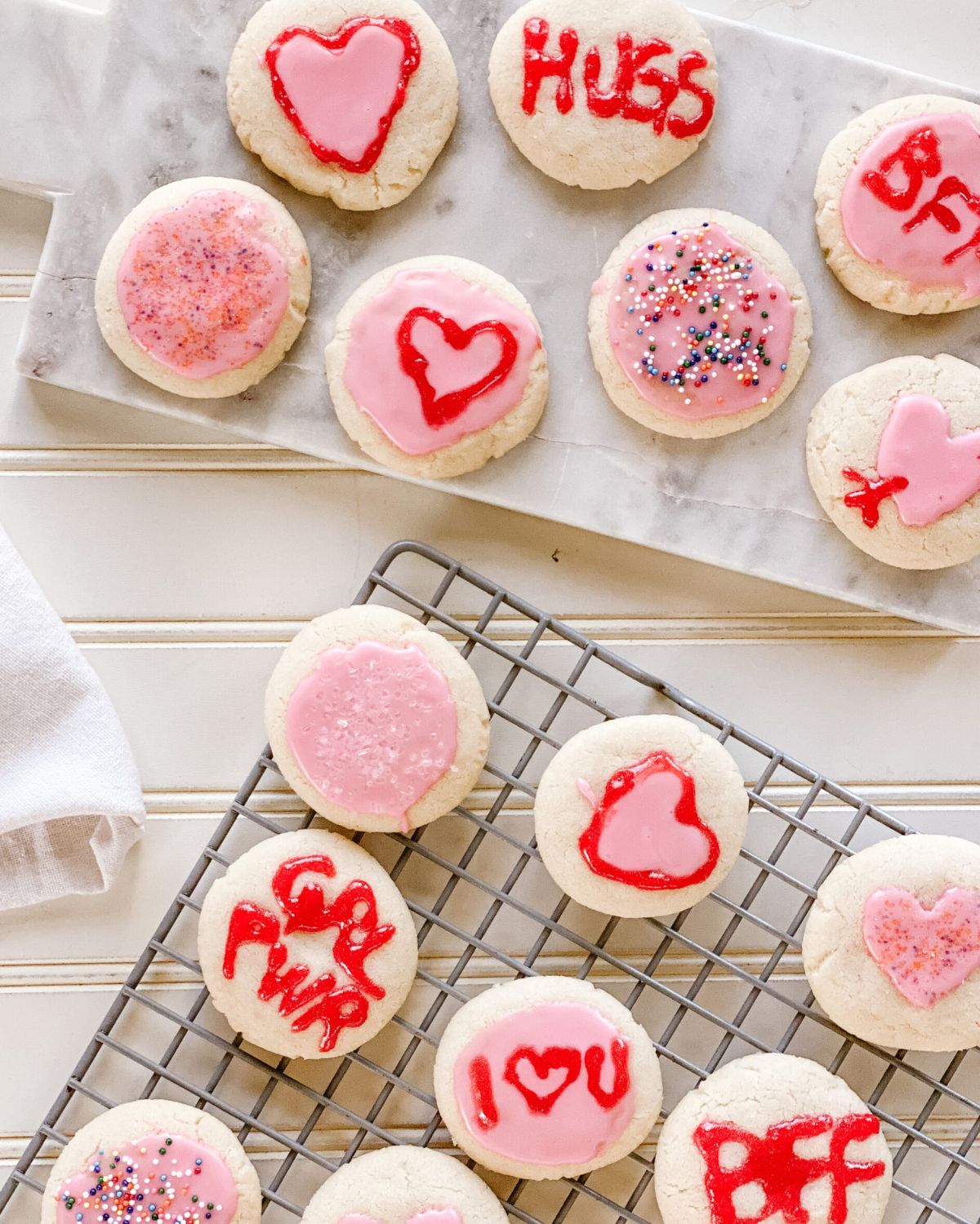 almond cookies with pink frosting and Valentine's Day messages on a marble board and wire cooling rack