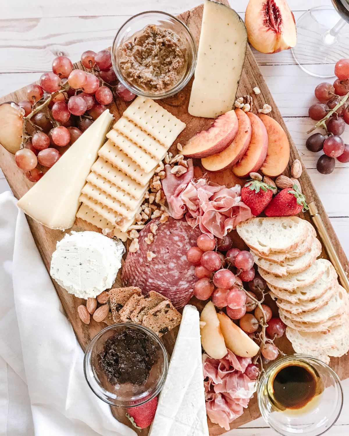 Charcuterie board with brie, aged gouda, herbed goat cheese, salami, prosciutto, olive tapenade, honey, fig jam, grapes, sliced peaches, strawberries, almonds, crackers, and sliced baguette