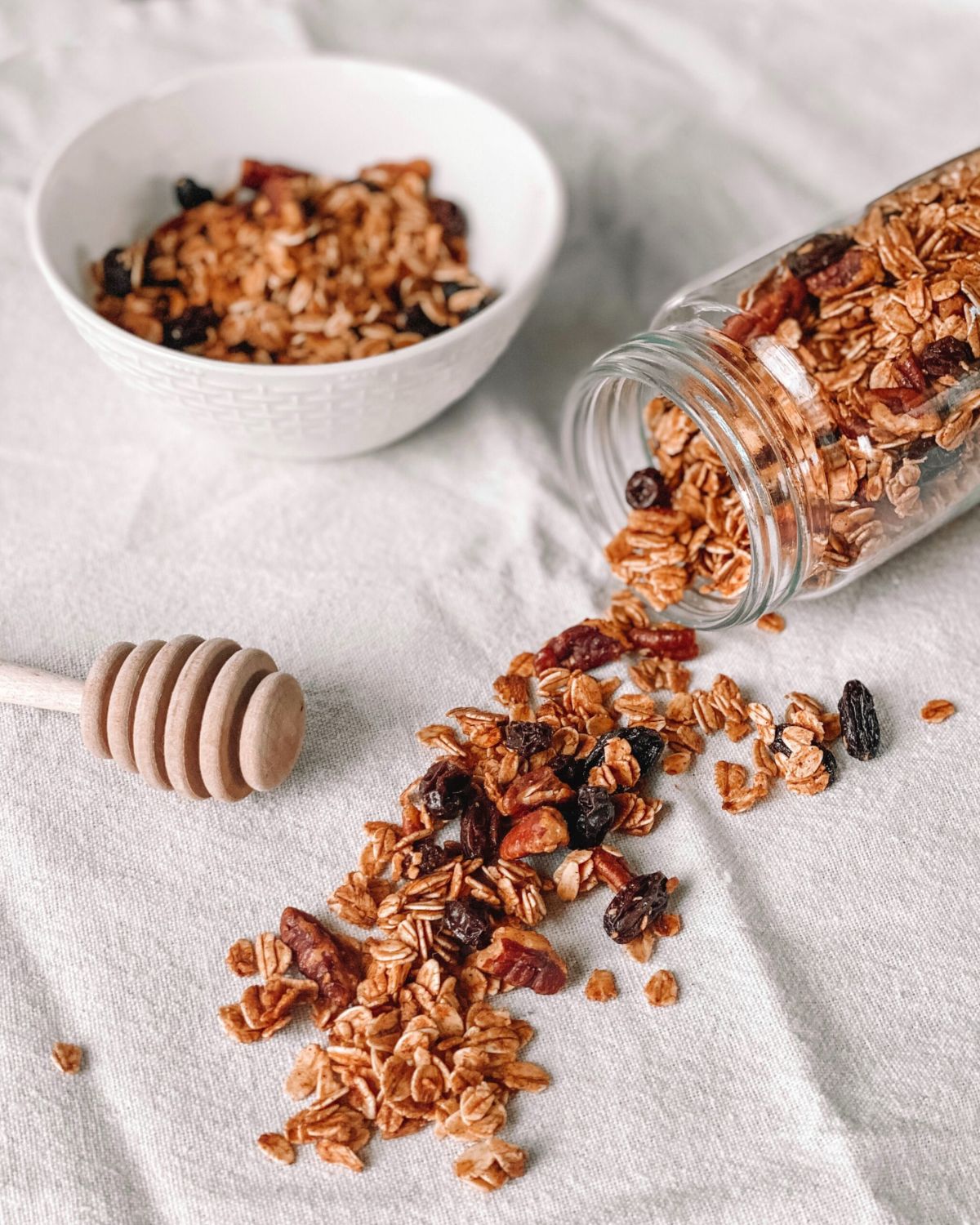 pumpkin spice granola spilling from a jar next to a white bowl of granola on a linen cloth
