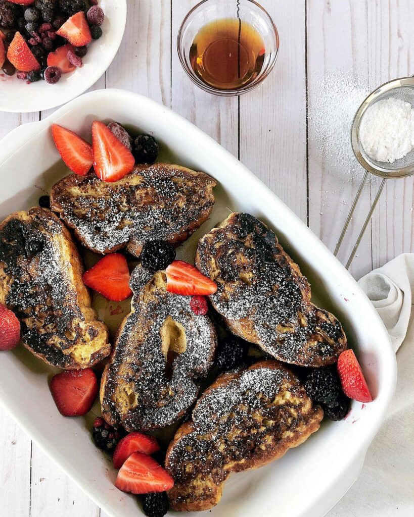Vanilla rum french toast in a white baking dish with fresh strawberries, blueberries, and powdered sugar