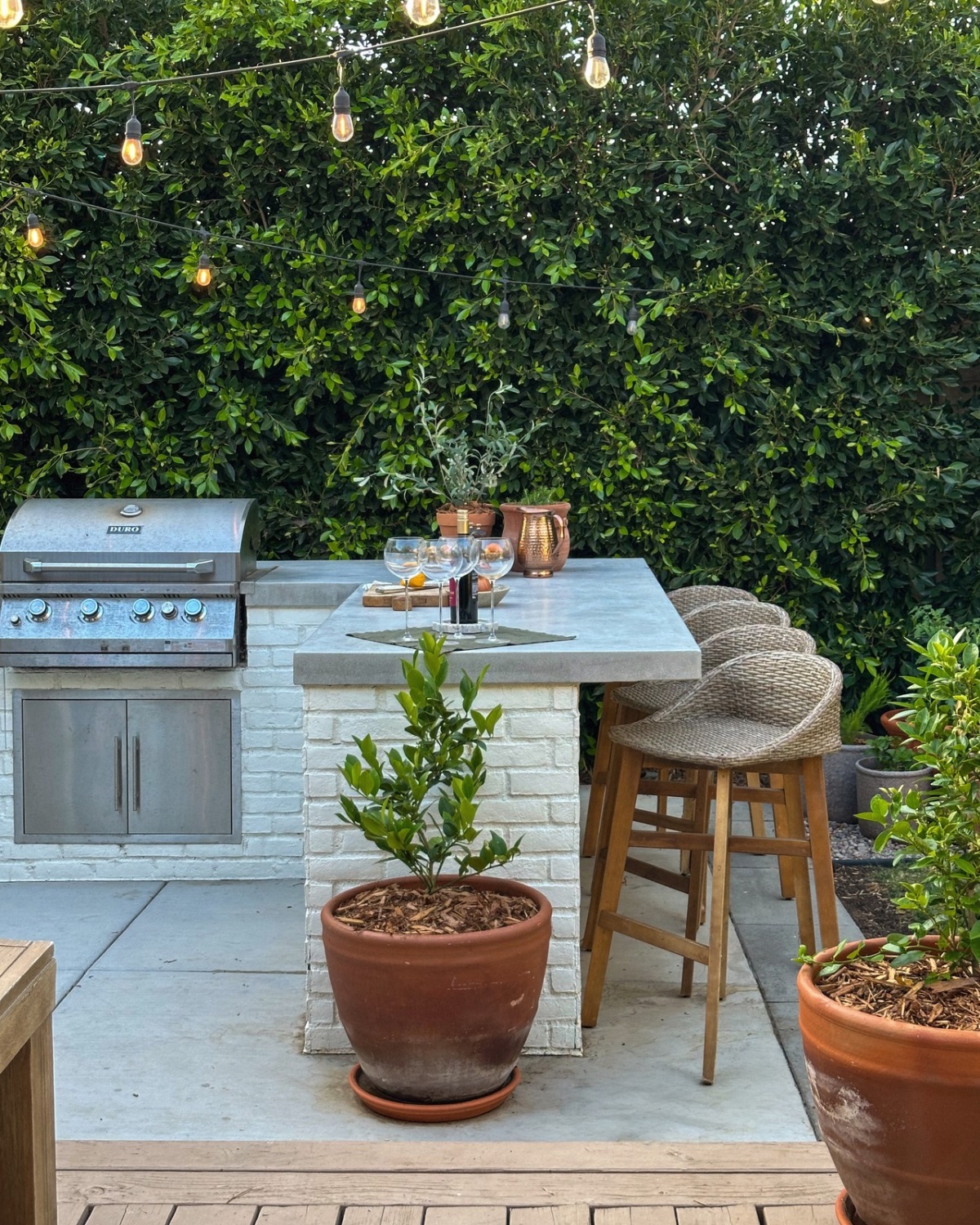 Built-in stainless steel grill and white brick bar with a concrete countertop, woven bar stools, terracotta planters and string lights
