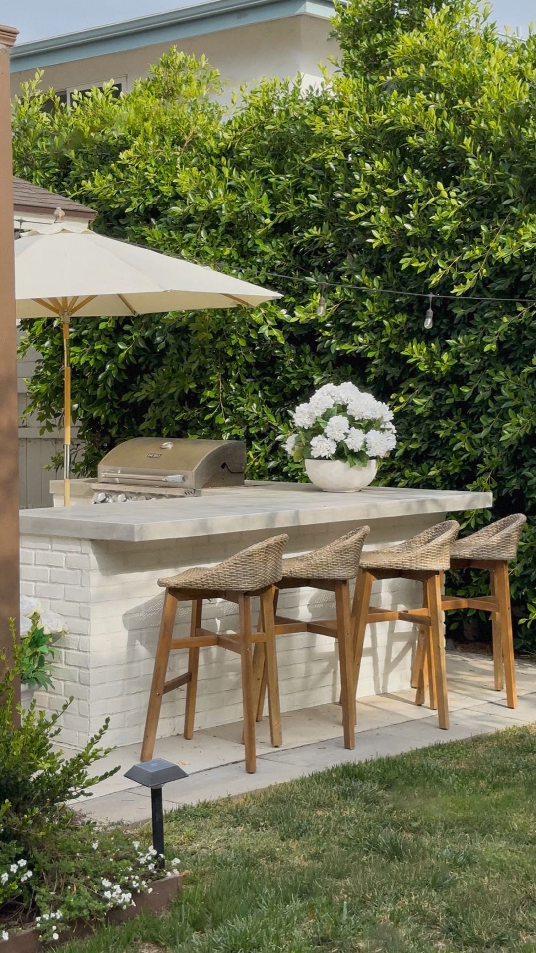 Four woven bar stools along a white brick outdoor bar with a concrete countertop, stainless grill and cream patio umbrella
