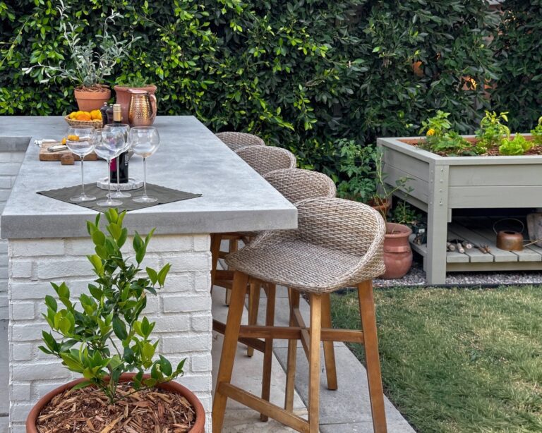 White brick outdoor bar with a concrete countertop, woven stools, wine glasses, and a raised garden bed against a lush privacy hedge.