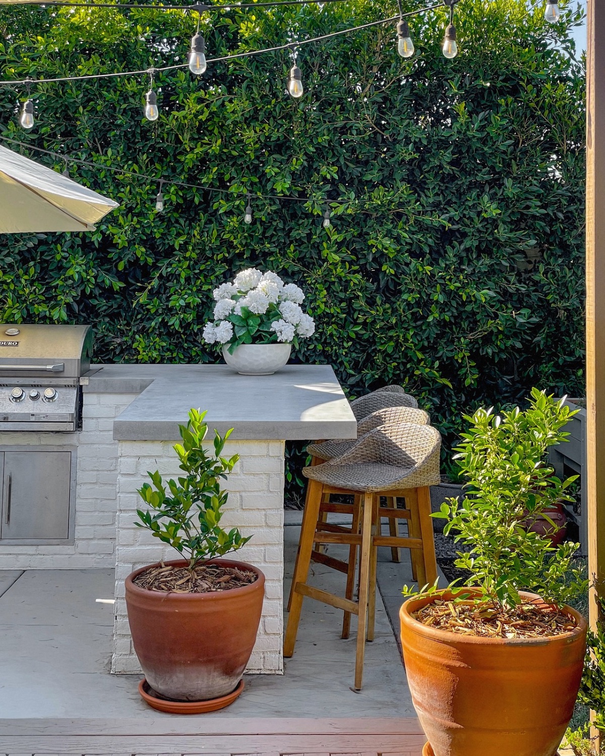 White brick outdoor bar with a concrete countertop topped with white hydrangeas, woven bar stools and string lights