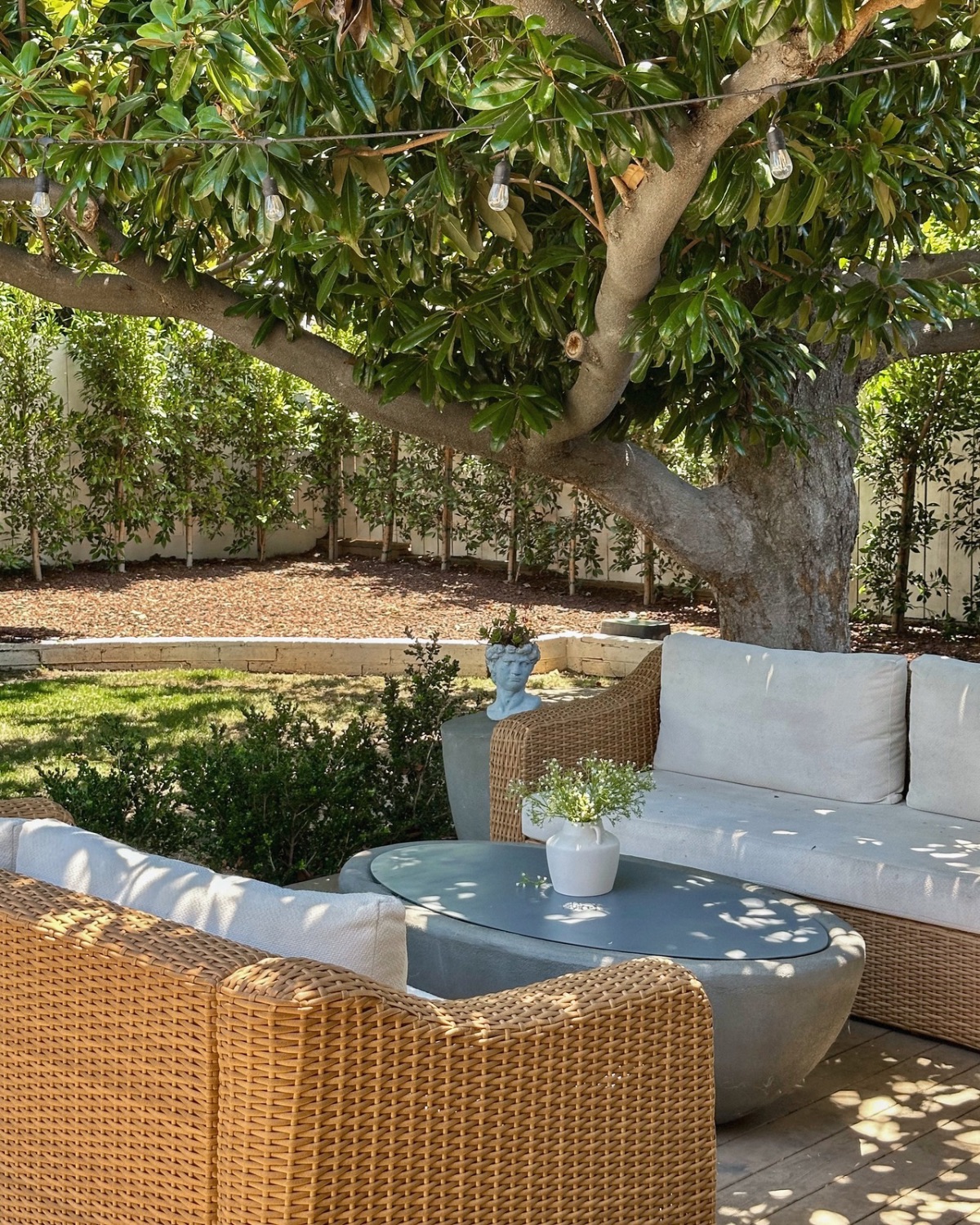 Wicker outdoor sofas with cream cushions around a round fire pit with a matching lid used as a coffee table, under a magnolia tree with string lights