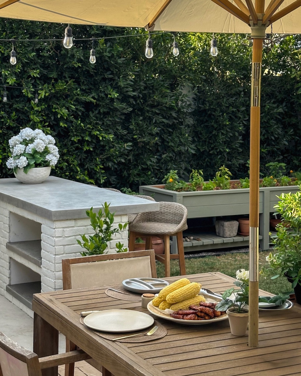 Wood outdoor dining table set with grilled chicken and corn on the cob under an ivory umbrella and market lights, with the raised garden behind
