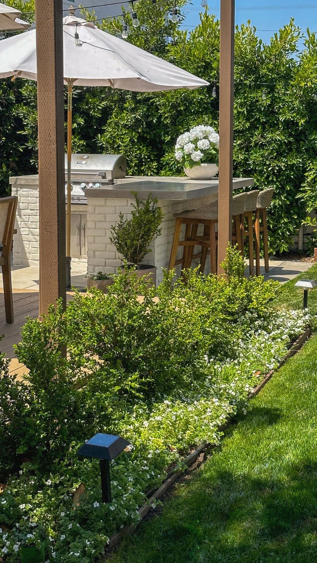 Garden bed of boxwoods and white bacopa with a lawn light along a fescue lawn, leading to the white brick outdoor kitchen