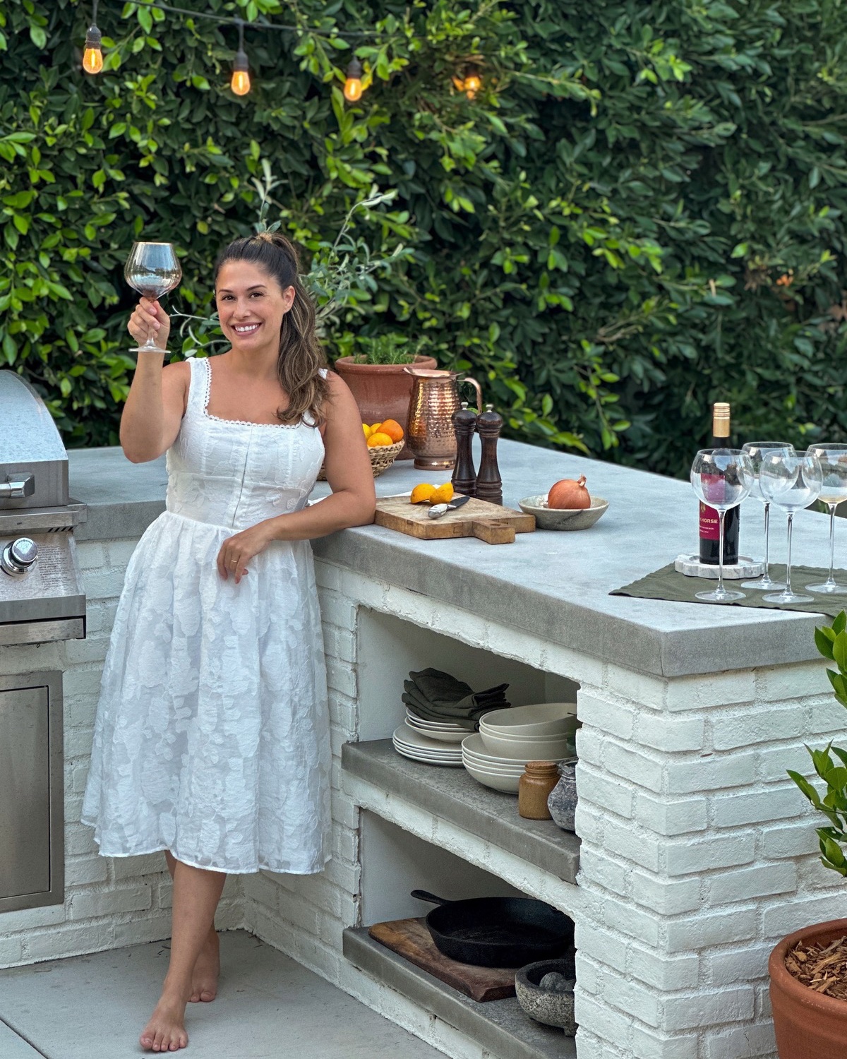 Liz Lovery raising a wine glass beside her white brick outdoor kitchen with concrete countertops and string lights