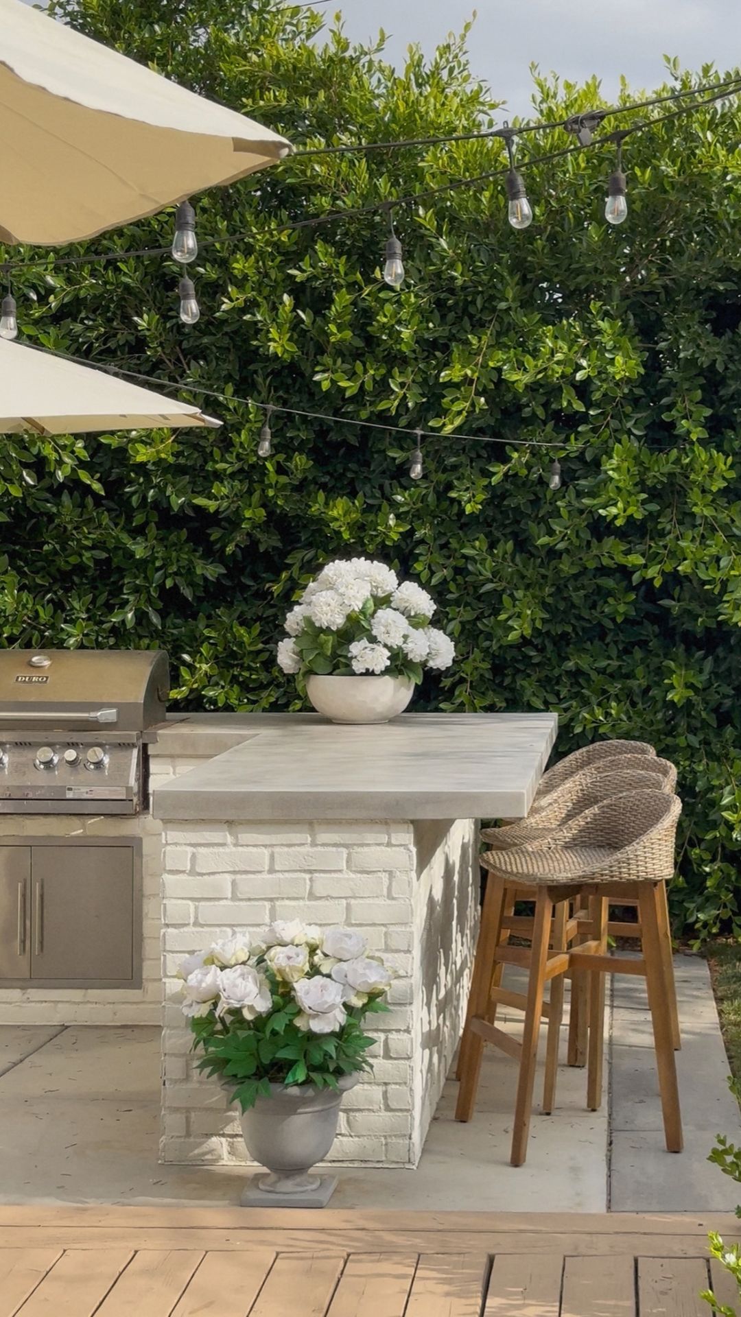 White brick outdoor bar with gray concrete countertops, woven wood bar stools, white flowers in urns and string lights