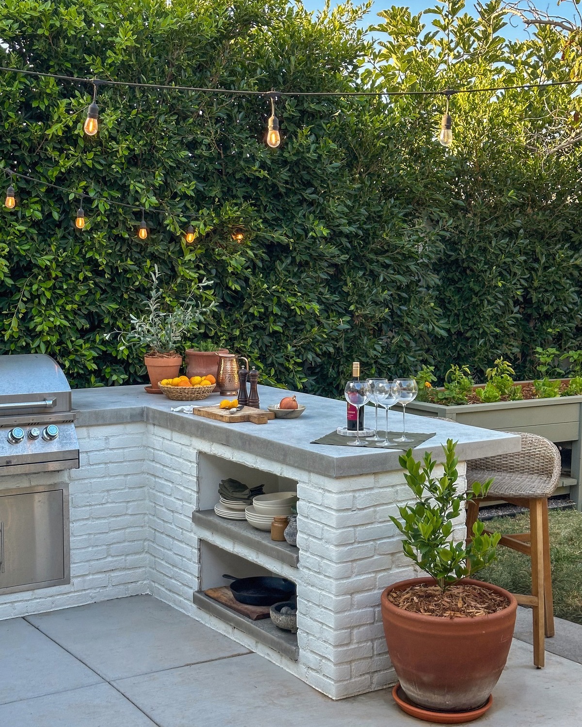 White painted brick outdoor kitchen with open shelving holding plates and a cast iron pan under a concrete countertop